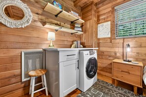 Ein kleiner Hauswirtschaftsraum mit Waschmaschine, Hocker, Tisch mit Lampe und Regal mit Büchern und einer Kiste.