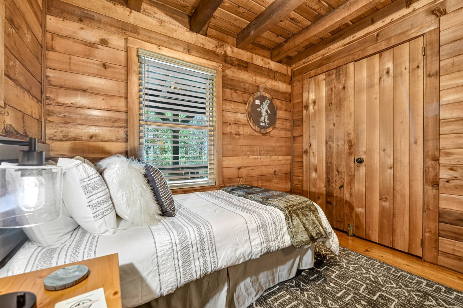 Ein Schlafzimmer einer Holzhütte mit einem Bett, einem Fenster mit Jalousien, einer Tür und einer an der Wand befestigten Uhr.
