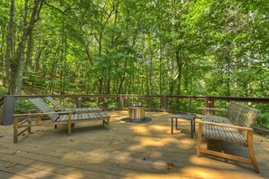 Une terrasse extérieure avec un plancher en bois, entourée d’arbres, comprenant des chaises longues et une table ronde.