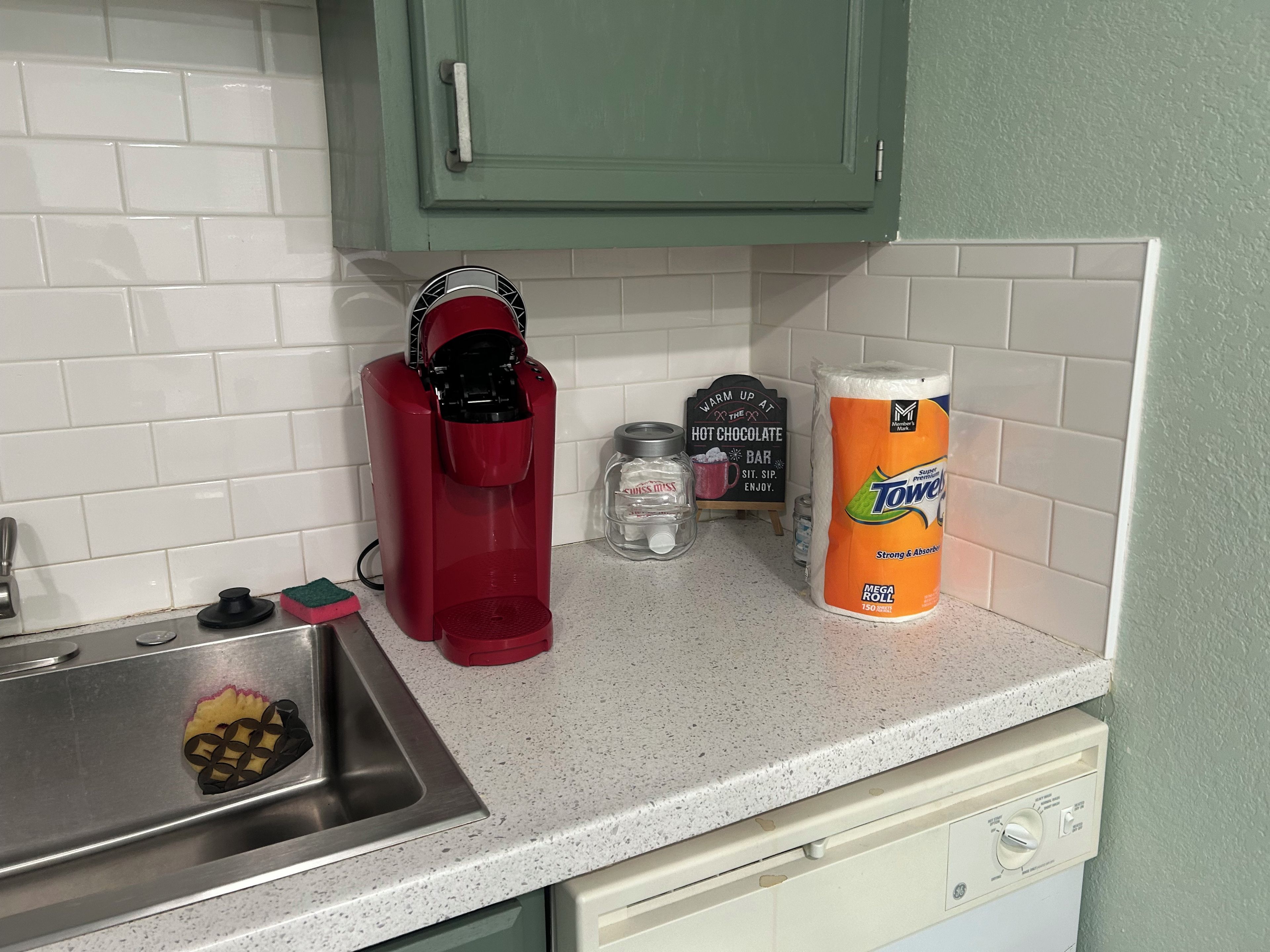 A kitchen with a coffee maker, a roll of paper towels, and a sign that says 'WARM UP AT HOT CHOCOLATE BAR.'