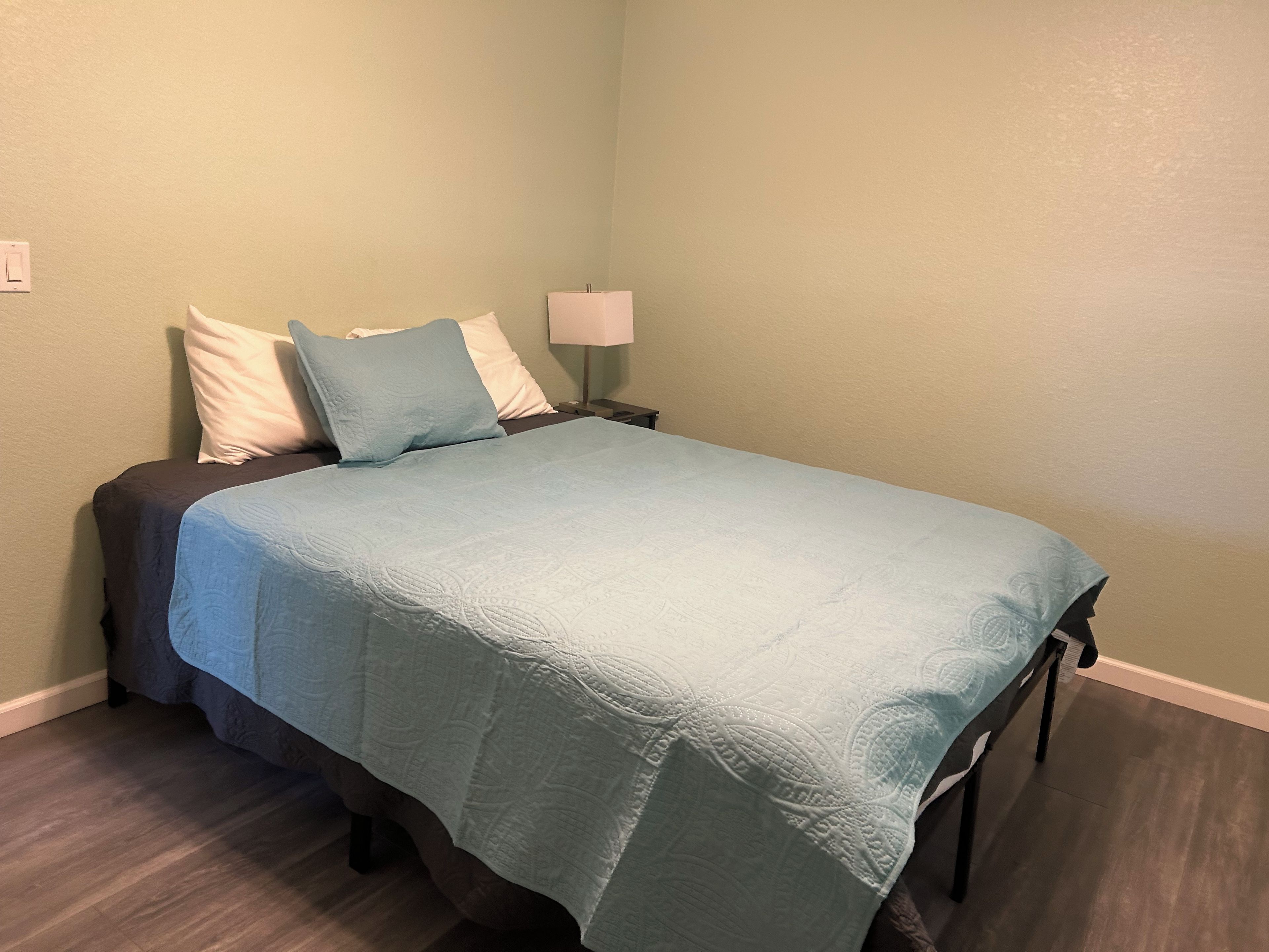 Een bed met een blauwe spreidek en kussens, een nachtkastje met een lamp, en een lichtschakelaar aan de muur.