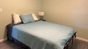 Een bed met een blauwe spreidek en kussens, een nachtkastje met een lamp, en een lichtschakelaar aan de muur.