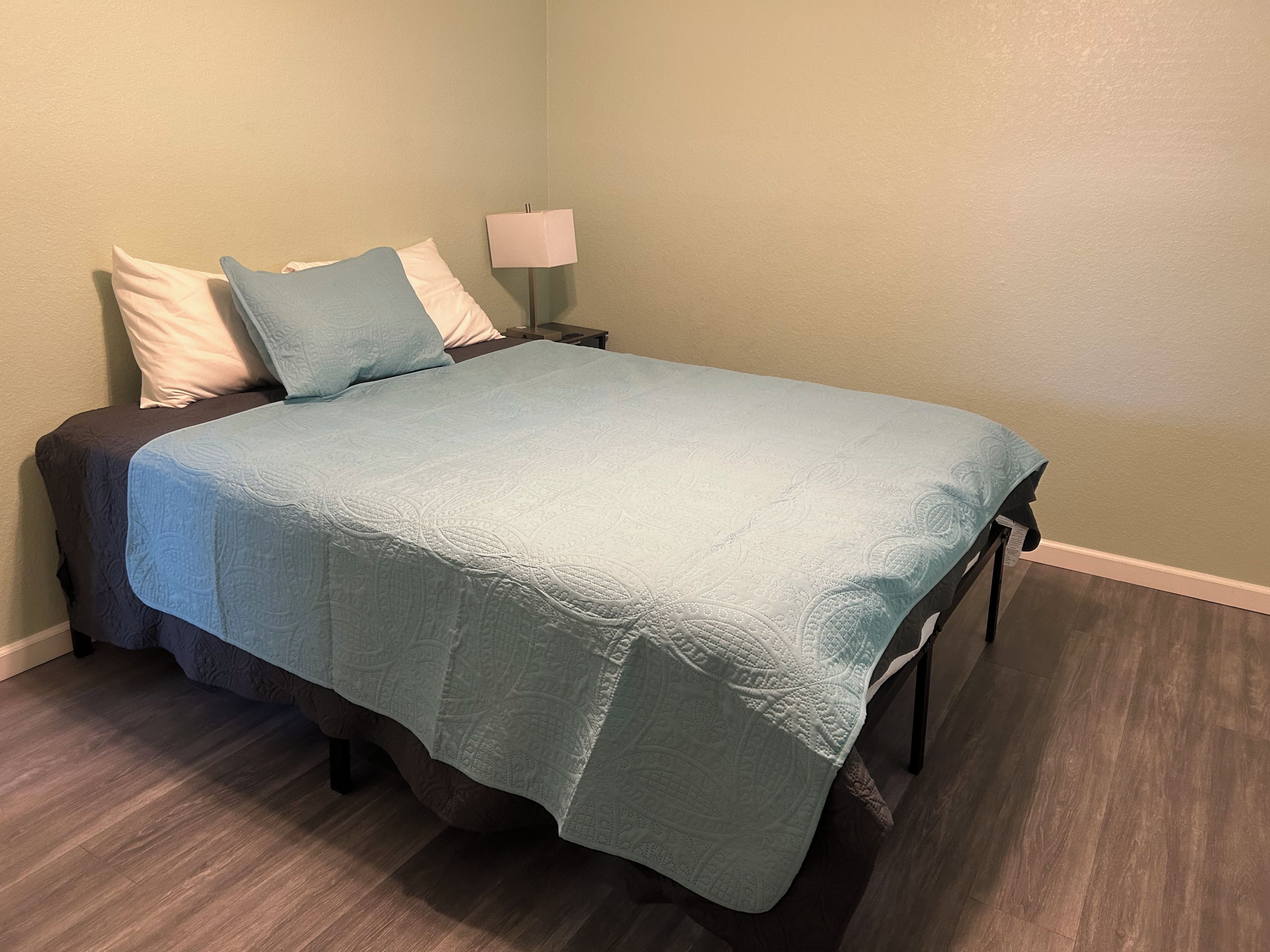 Una cama con colcha azul, una lámpara de noche y una mesita.