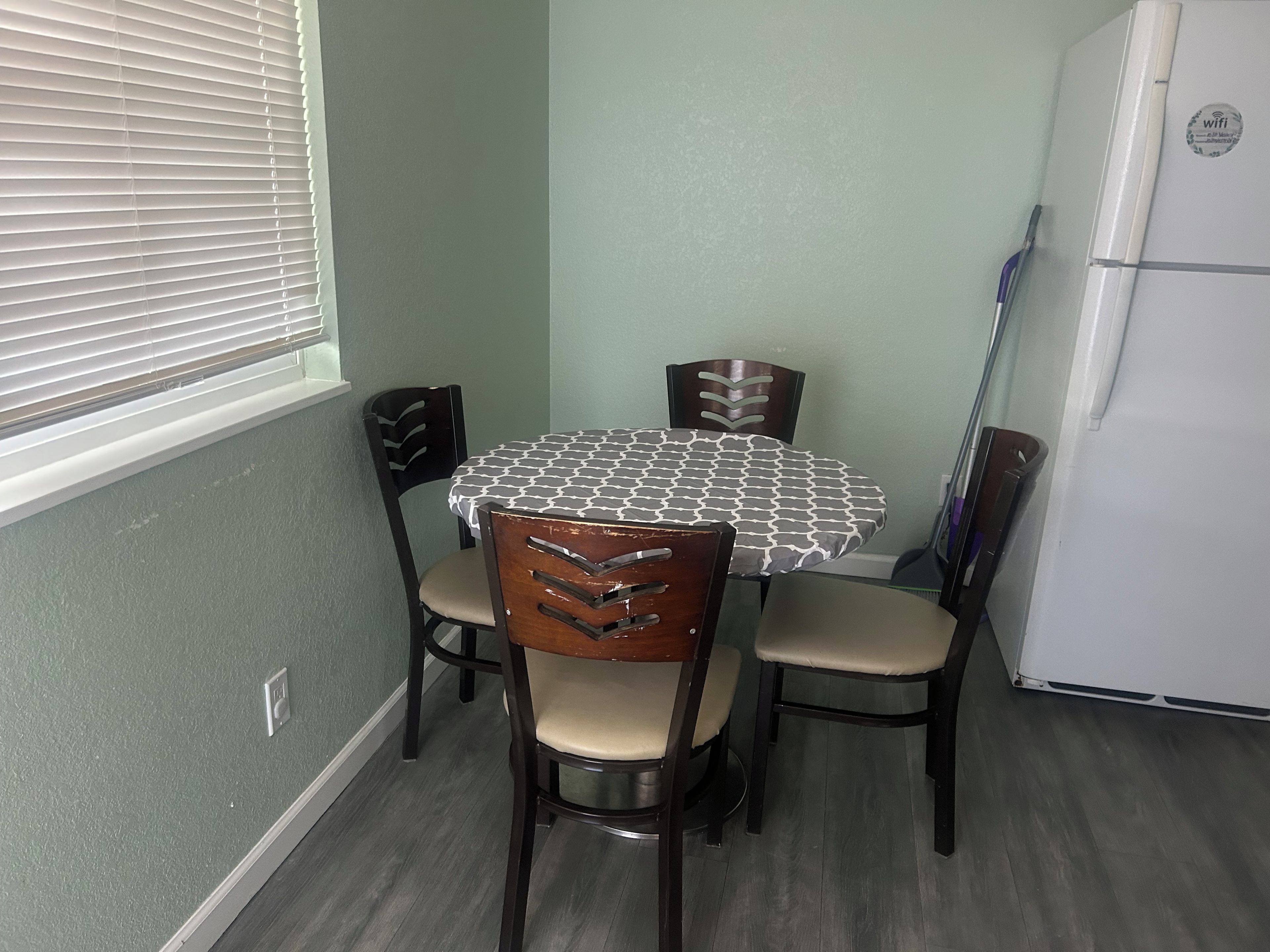 A dining area with a round table, four chairs, a window with blinds, and a refrigerator.