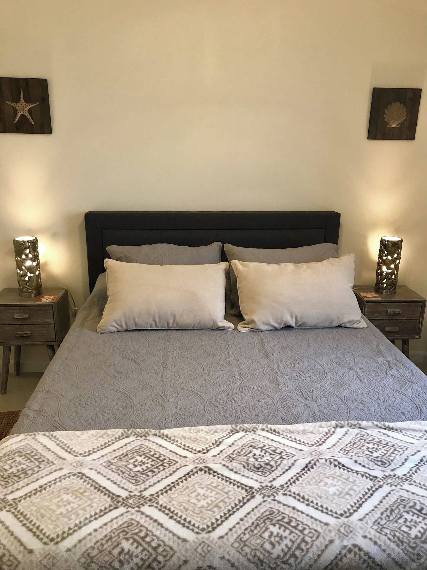 Een bed met een donker hoofdbord, twee kussens en een beddegoed met dessin. Twee nachtkastjes met decoratieve lampen staan ernaast.