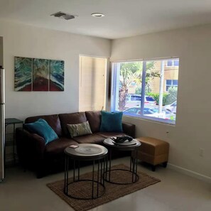 A living room with a brown leather sofa, two round tables, a rug, and a window with blinds. - Sun & Sand Retreat 1BR 1BA (Hollywood)