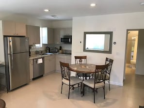 Cozinha moderna com eletrodomésticos de aço inoxidável, área de jantar com mesa redonda e cadeiras, e vista para o exterior através de uma janela.