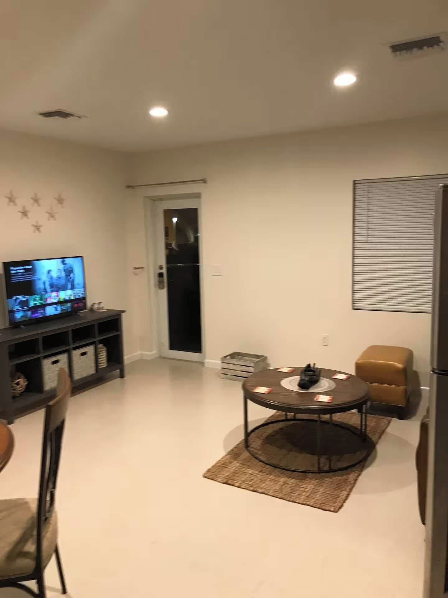 Una sala de estar moderna con una televisión de pantalla plana, una mesa de comedor con sillas, una mesa de centro y un refrigerador.