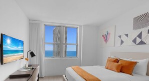 A hotel room with a bed, a desk, a window with a view of a tall building, and artwork on the walls. - Modern 2BD 2BA Resort Luxury Bay Corner Views (Hollywood)