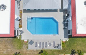 Eine Vogelperspektive zeigt einen Swimmingpool mit einer Liegefläche, umgeben von einem Gebäude mit rotem Dach und einer überdachten Terrasse.