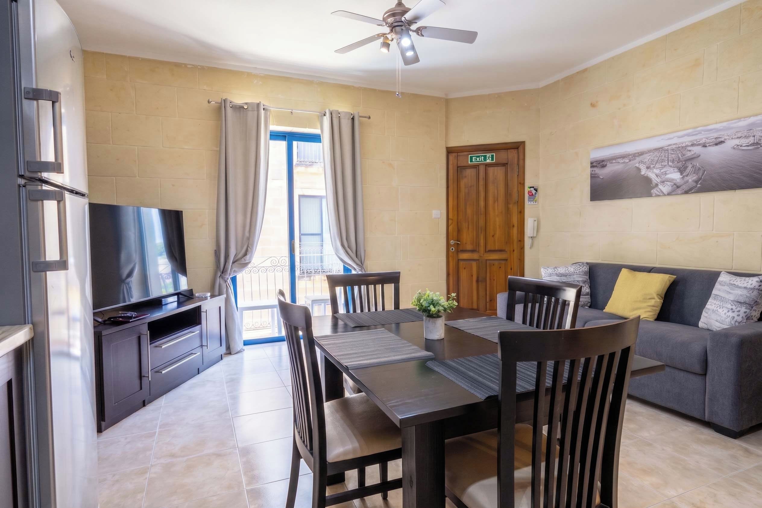 Una habitación con una mesa de comedor, sillas, un televisor, un ventilador de techo y una puerta con la indicación 'Exit'.