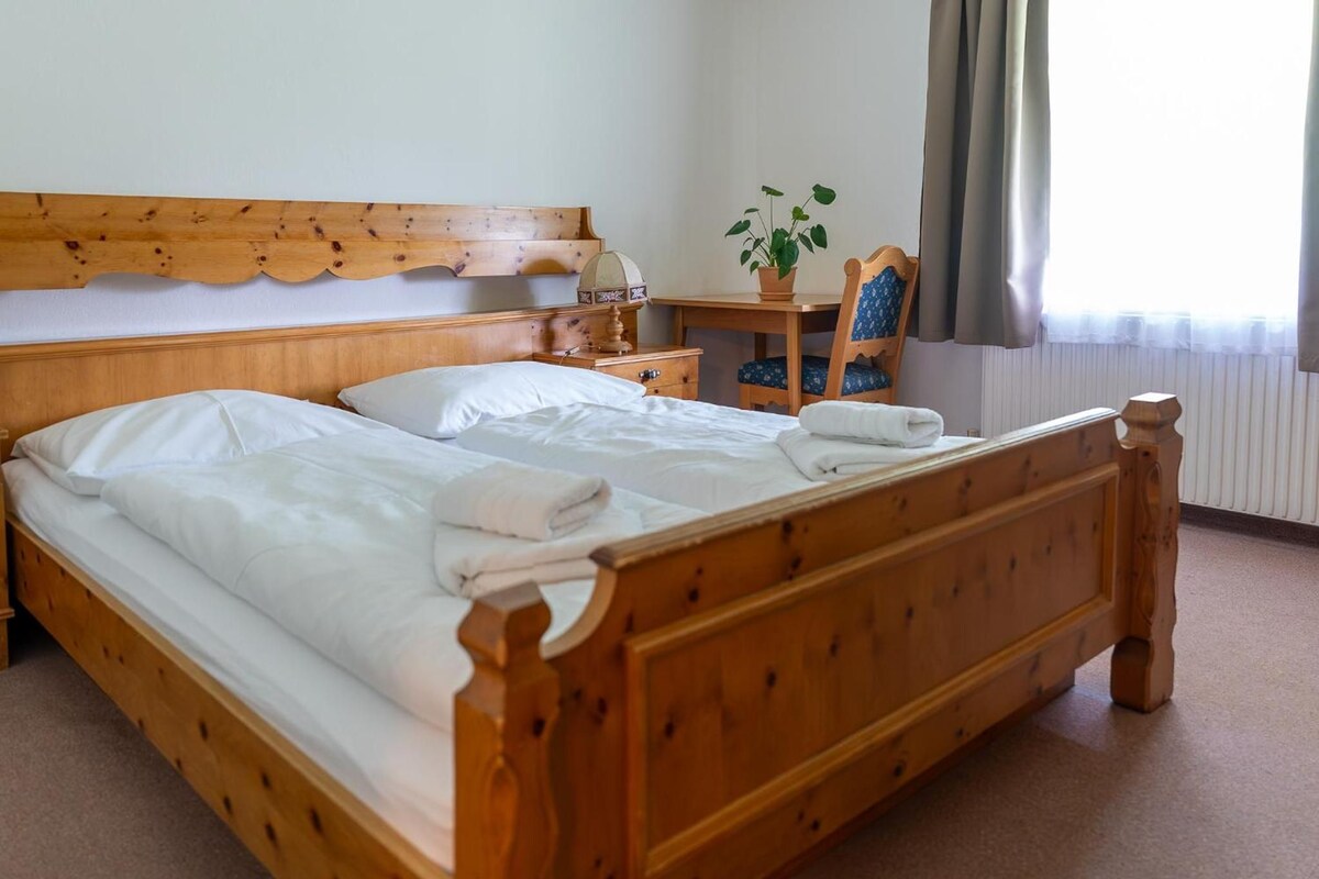 A double bed with a wooden headboard and footboard, a bedside table with a lamp, a chair with a cushion, and a potted plant.