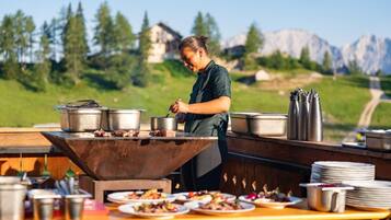Ein Koch bereitet Essen im Freien zu, mit Blick auf die Berge im Hintergrund.