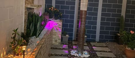 Ein Außenbereich bei Nacht mit einer Ziegelwand, Lichterketten, einem Palmenbaum und Blumentöpfen.