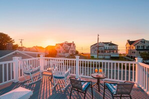 A deck with white railings, a round table with chairs, and a sunset view of houses. - Moody Surfer - Coastal Retreat (Wells)