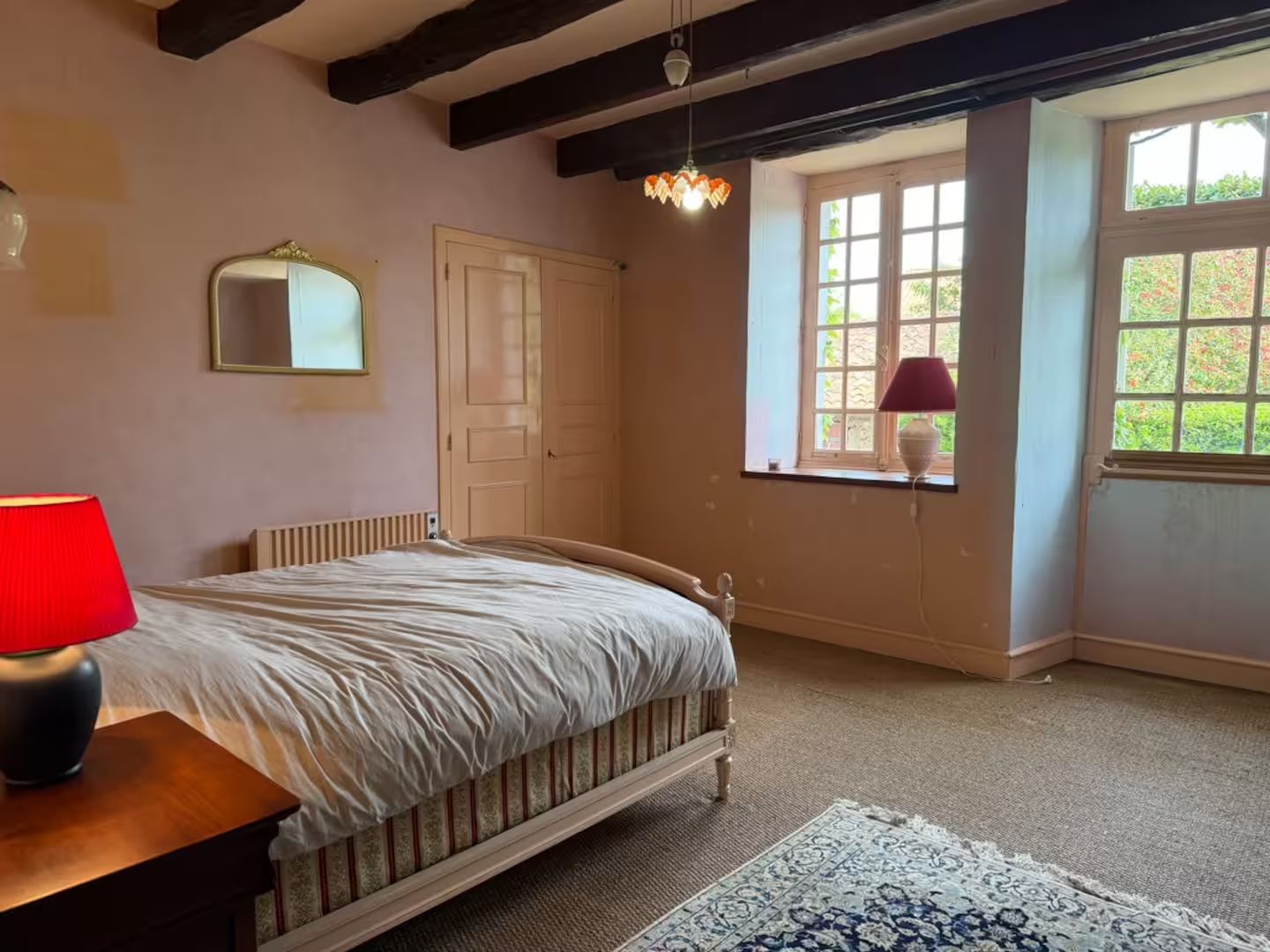 A bedroom with a bed, a bedside table with a lamp, a window with a view, and a hanging light fixture.