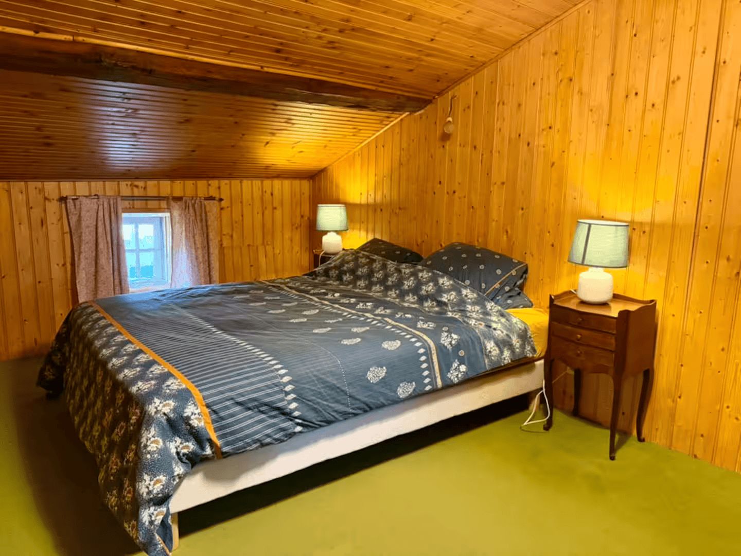 Ein Zimmer mit Holzwänden, einem Bett, zwei Nachttischlampen und einem mit Vorhängen drapierten Fenster.