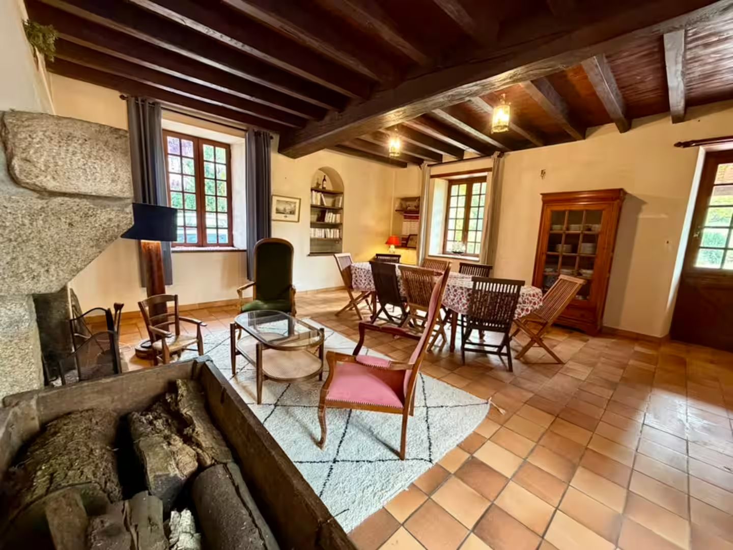 Una habitación acogedora con chimenea, vigas de madera, un comedor con sillas y una mesa, y una estantería.