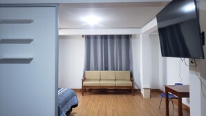 Huoneessa on sohva, seinälle asennettu televisio, puulattia ja valkoinen ovi.