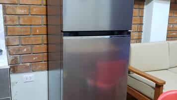 A stainless steel refrigerator with a control panel and a lower compartment, placed on a wooden floor against a brick wall.