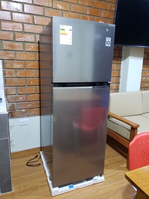 A stainless steel refrigerator with a control panel and a lower compartment, placed on a wooden floor against a brick wall.
