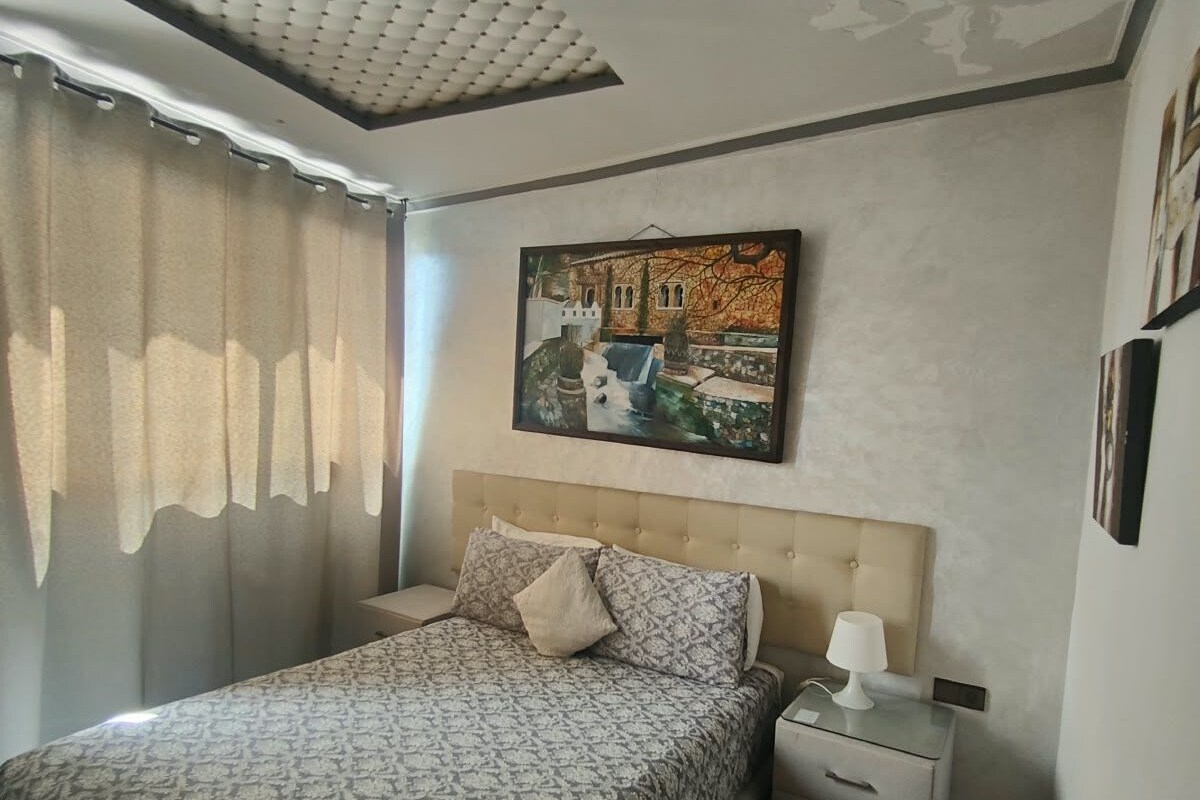 A bedroom with a bed, a nightstand, a lamp, a painting, and a ceiling with a textured pattern.