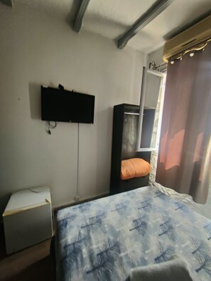 A bedroom with a bed, a small desk, a television mounted on the wall, and a window with curtains.