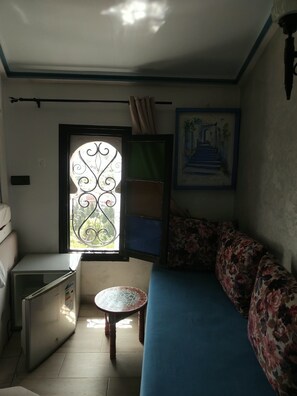 A compact living space with a sofa, a small table, a refrigerator, a window with decorative ironwork, and a framed painting on the wall.