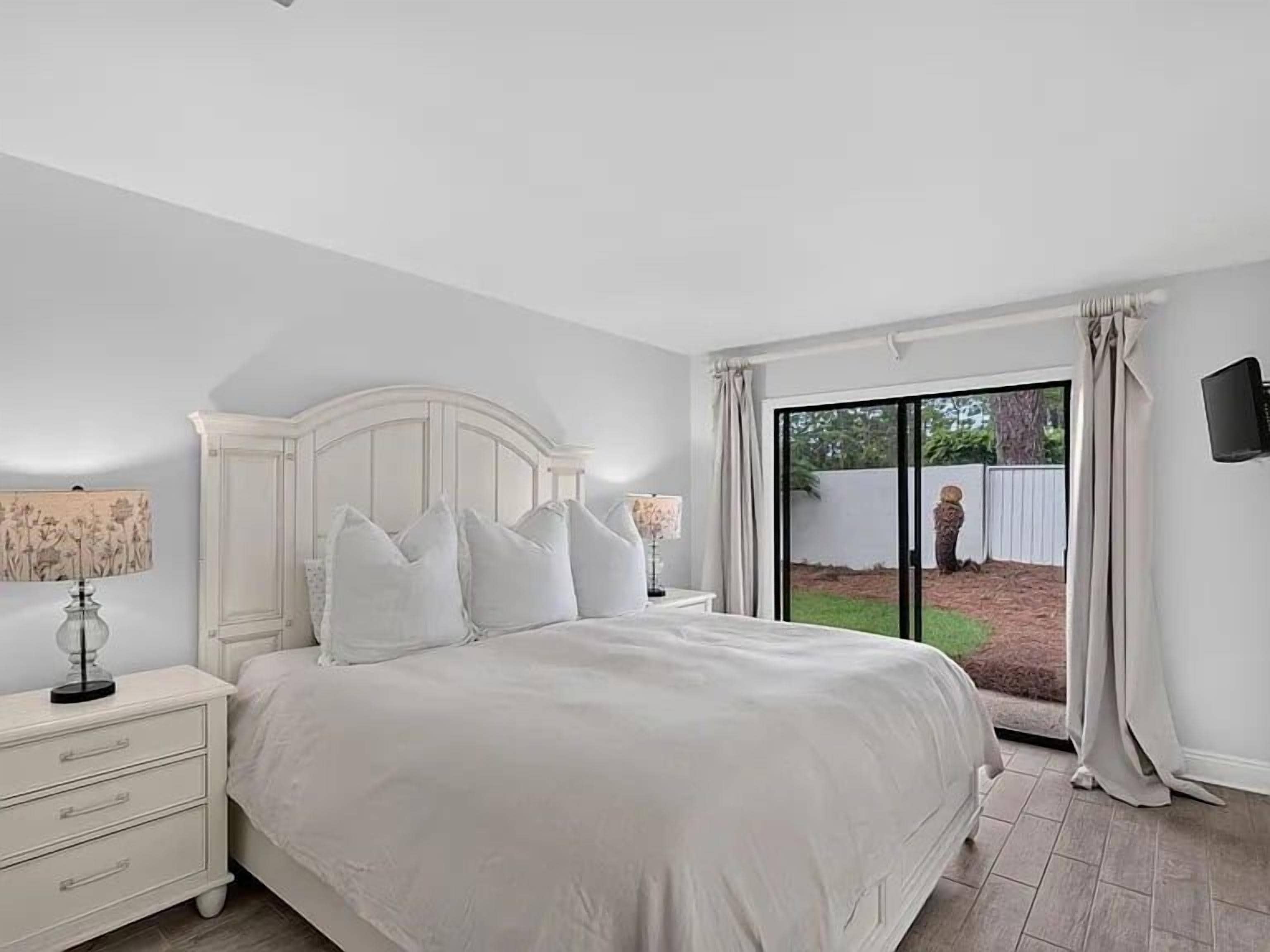 A bedroom with a large bed, a nightstand, a lamp, and a view of the outdoors through a sliding glass door.