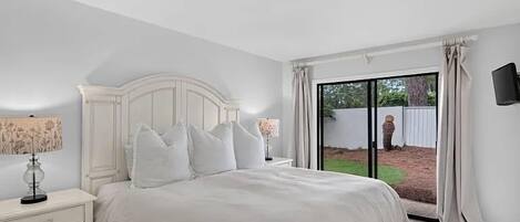 A bedroom with a large bed, a nightstand, a lamp, and a view of the outdoors through a sliding glass door.
