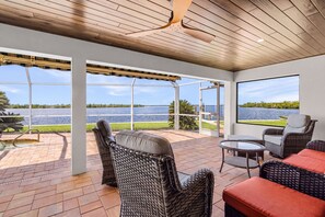 Eine überdachte Terrasse mit Korbmöbeln, einem Glastisch und Blick auf einen See.