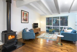 A living room with a wood-burning stove, blue sofas, a rug, and a large window with a view of trees. - Belle Cottage (Montrose)