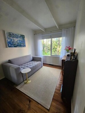 A living room with a grey sofa, a small round table, a rug, a window with curtains, and a painting on the wall. - Belle Cottage (Montrose)