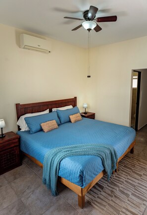 Een slaapkamer met een bed, nachtkastje, lamp, plafondventilator en airconditioning.