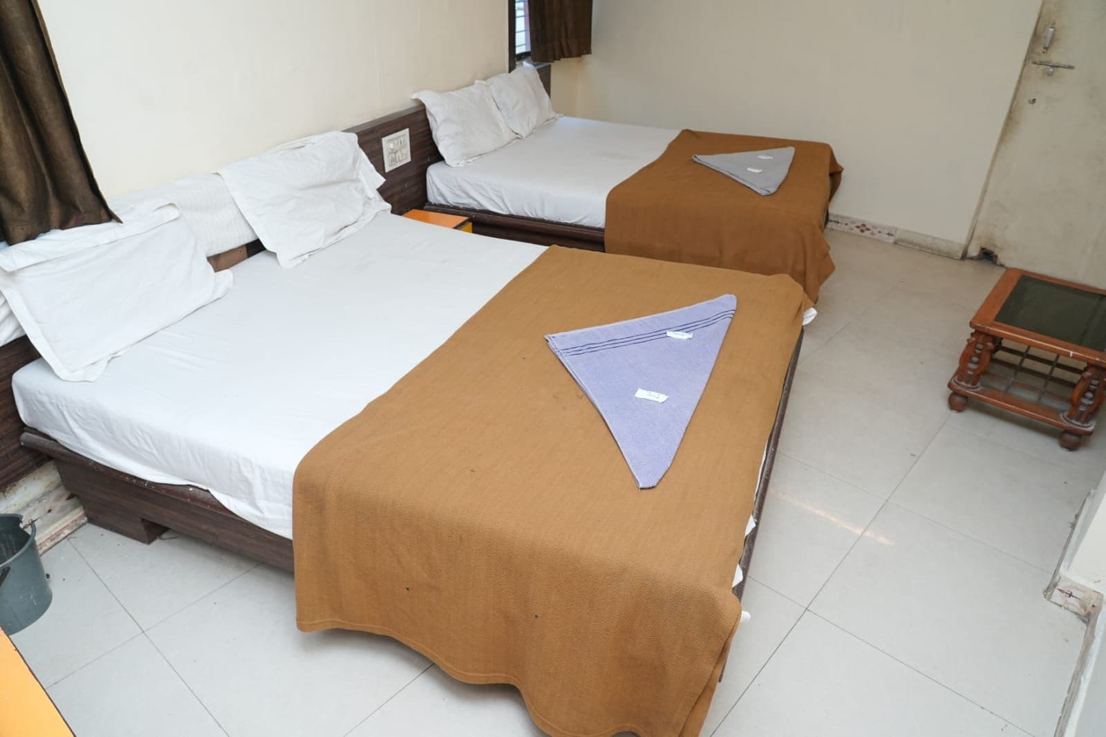 A room with two beds, each with a brown bedspread and white linens. There is a small wooden table with a glass top in the corner.