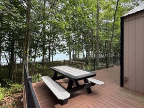 A wooden deck with a picnic table and benches, surrounded by trees and overlooking a body of water. - Fish Cabin – 1 Bedroom + Loft • Sleeps 6 • Lake & Beach Views • Access (La Pointe)