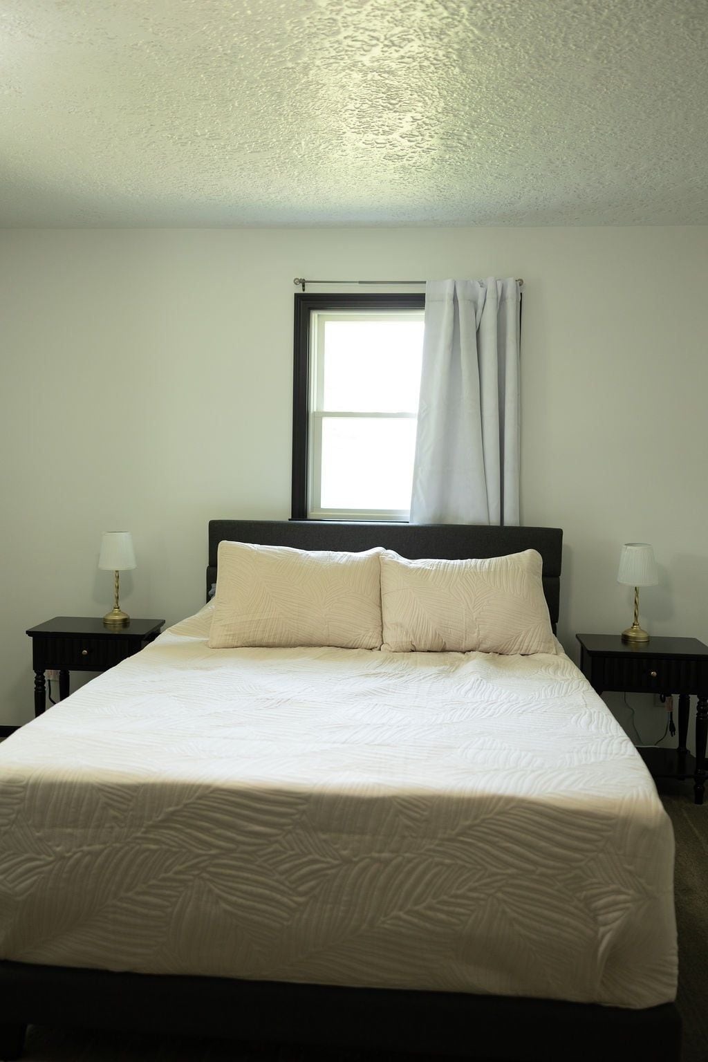 A bedroom with a bed, two bedside tables, a window with curtains, and a wall with a textured finish.