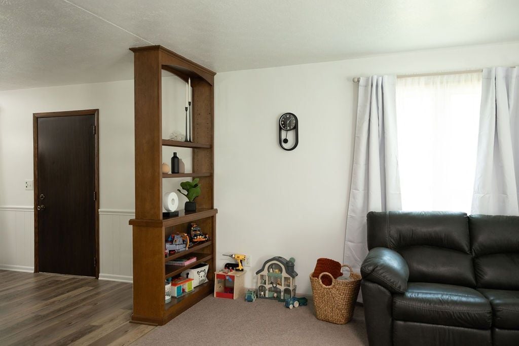 A living room with a dark brown wooden bookshelf, a black leather sofa, a wall clock, and a window with white curtains.