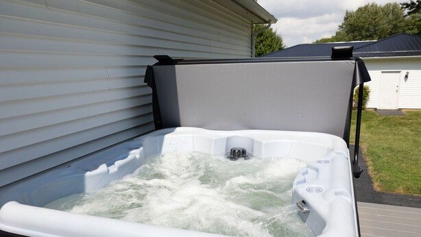 Ein Whirlpool mit weißer Außenverkleidung, draußen neben einem Haus mit weißer Fassade und dunklem Dach.