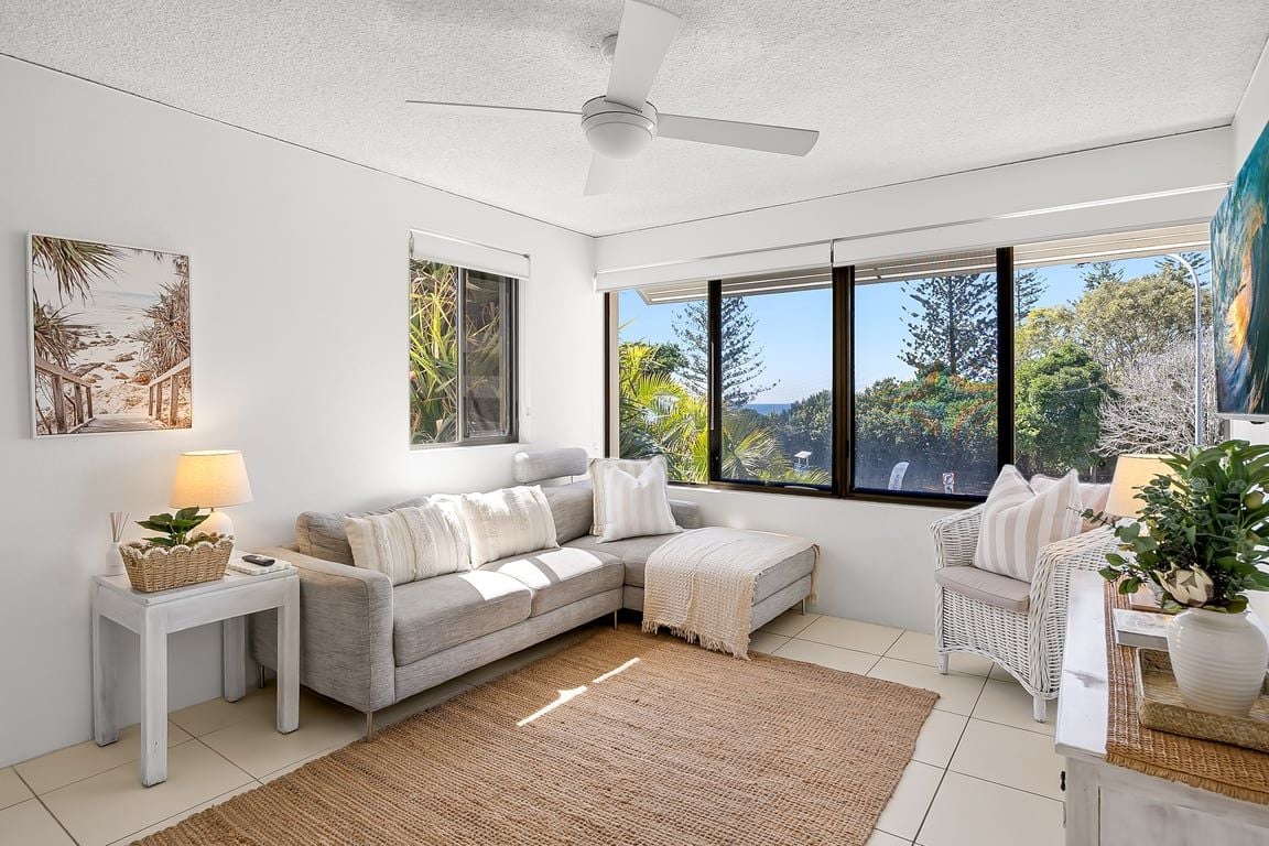 A modern living room with a grey sofa, a white side table, a large window with a view of greenery, and a ceiling fan.