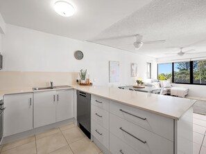 A modern kitchen with a central island, a built-in dishwasher, and a dining area with a sofa and table. - Beachside Views 2 Bedroom Poolside Apt Mooloolaba (Mooloolaba)