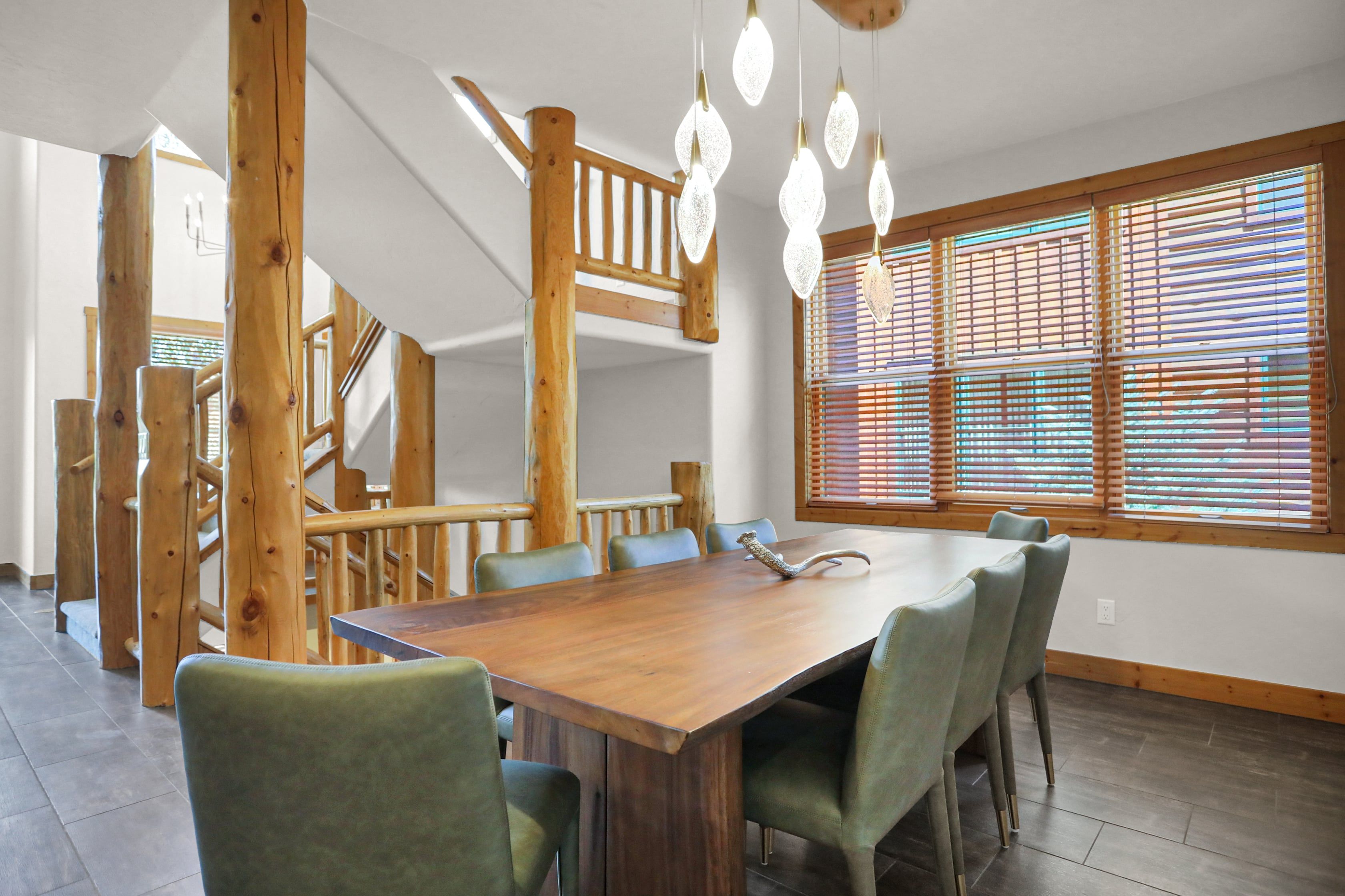 A dining area with a wooden table and chairs, a staircase, and large windows with blinds.