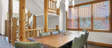 A dining area with a wooden table and chairs, a staircase, and large windows with blinds.