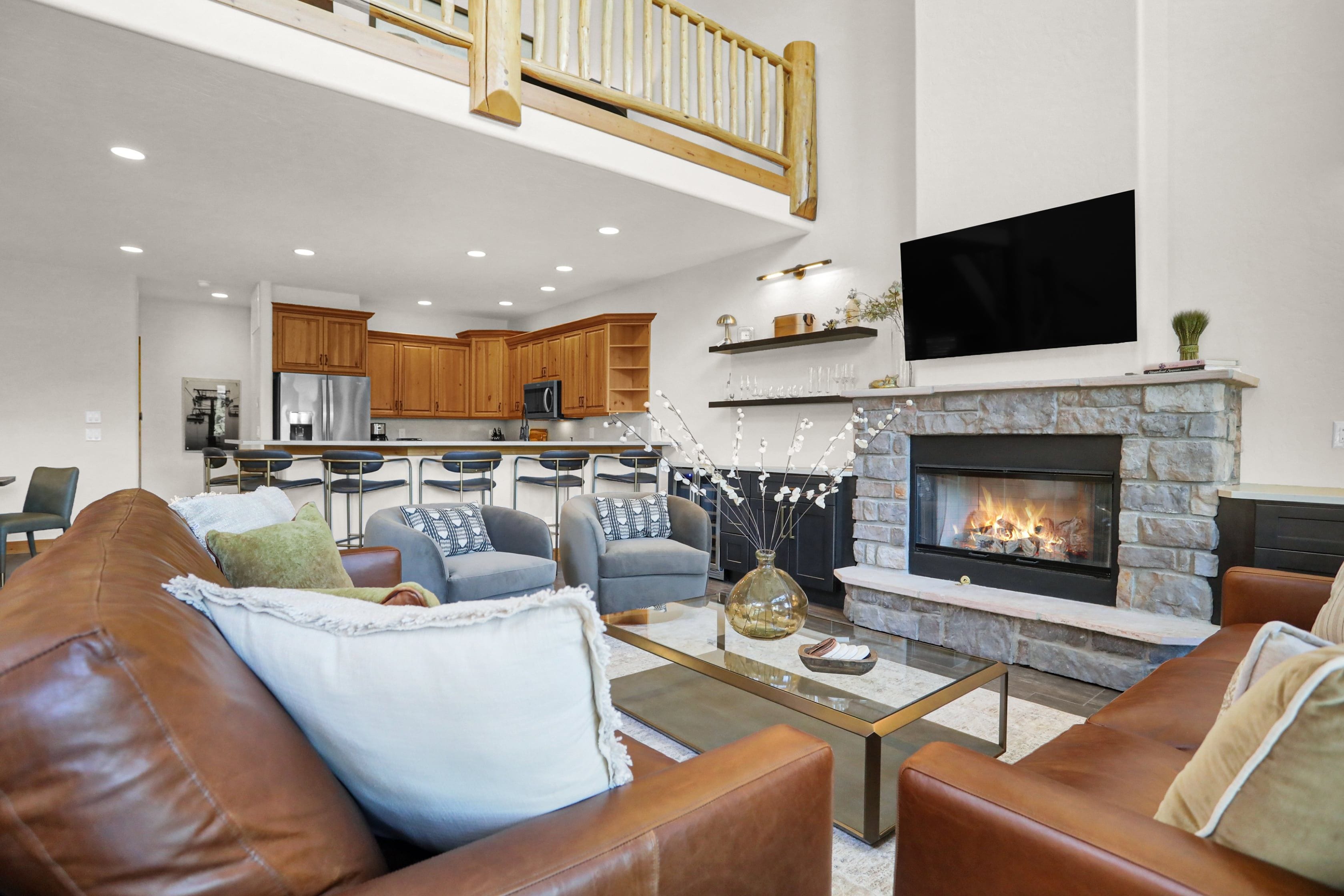 A modern living room with leather sofas, a glass coffee table, a fireplace, and a kitchen in the background.