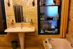 Casa de banho com lavatório, espelho e banheira. Há uma prateleira com objetos decorativos e um contentor de lixo.
