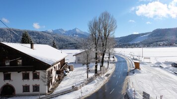 Una carretera cubierta de nieve que lleva a un edificio con techo inclinado, rodeado de árboles desnudos y montañas al fondo.