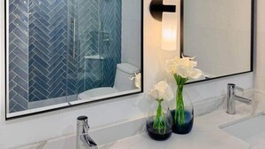 A bathroom with a large mirror, two sinks, and a shower with a herringbone tile pattern. - NEWLY Remodeled - Oceanfront Paradise (Honolulu)