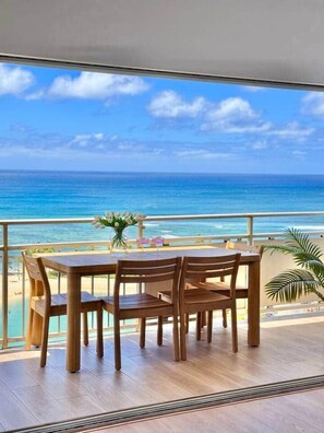 A balcony with a wooden table and chairs, overlooking a beach and ocean. - NEWLY Remodeled - Oceanfront Paradise (Honolulu)