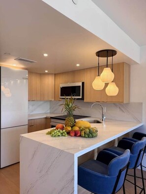 A modern kitchen with a marble island, wooden cabinets, a stainless steel sink, and a fruit bowl. - NEWLY Remodeled Oceanfront Fireworks Every Friday (Honolulu)