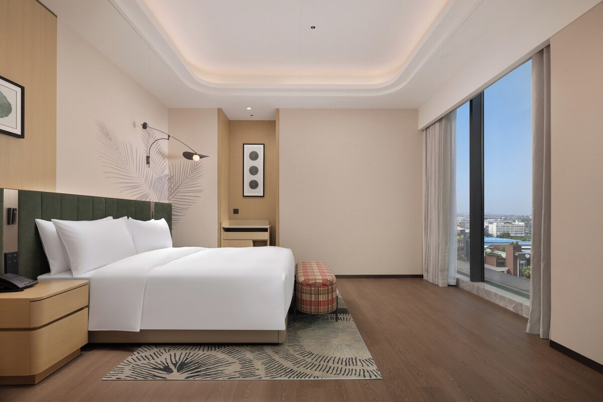 Habitació d'hotel moderna amb un llit gran, una petita taula de nit, un escriptori, una cadira, vista a la ciutat i una finestra ampla.